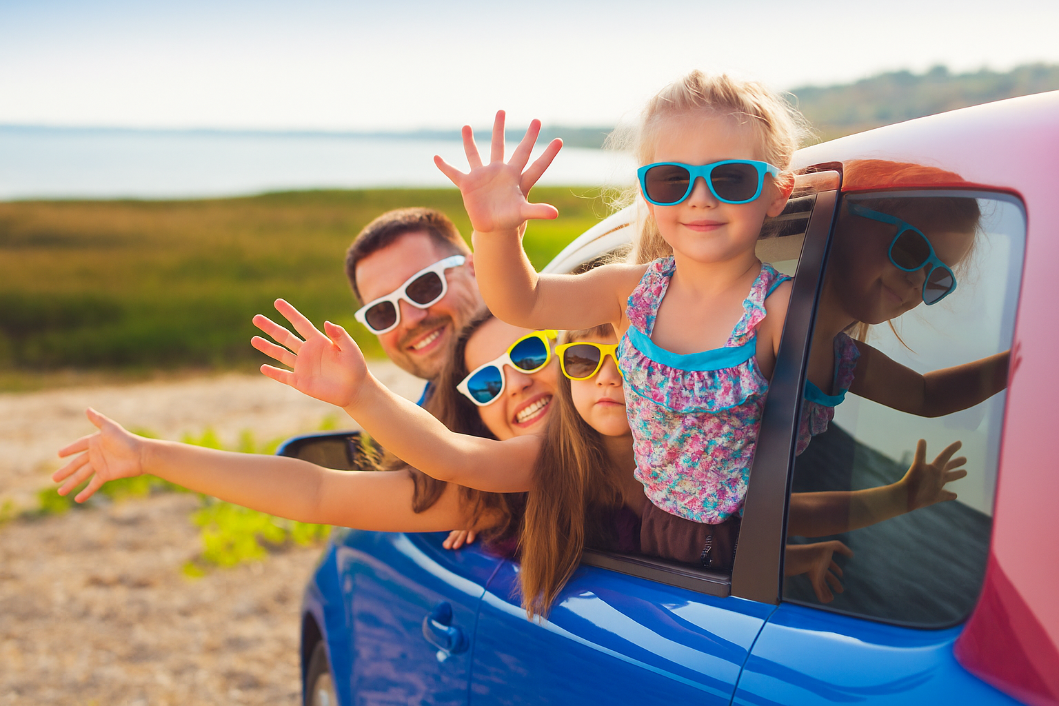Vater, Mutter und zwei Töchter winken aus dem Auto und stehen mit dem Wagen an einer sonnigen Küste.